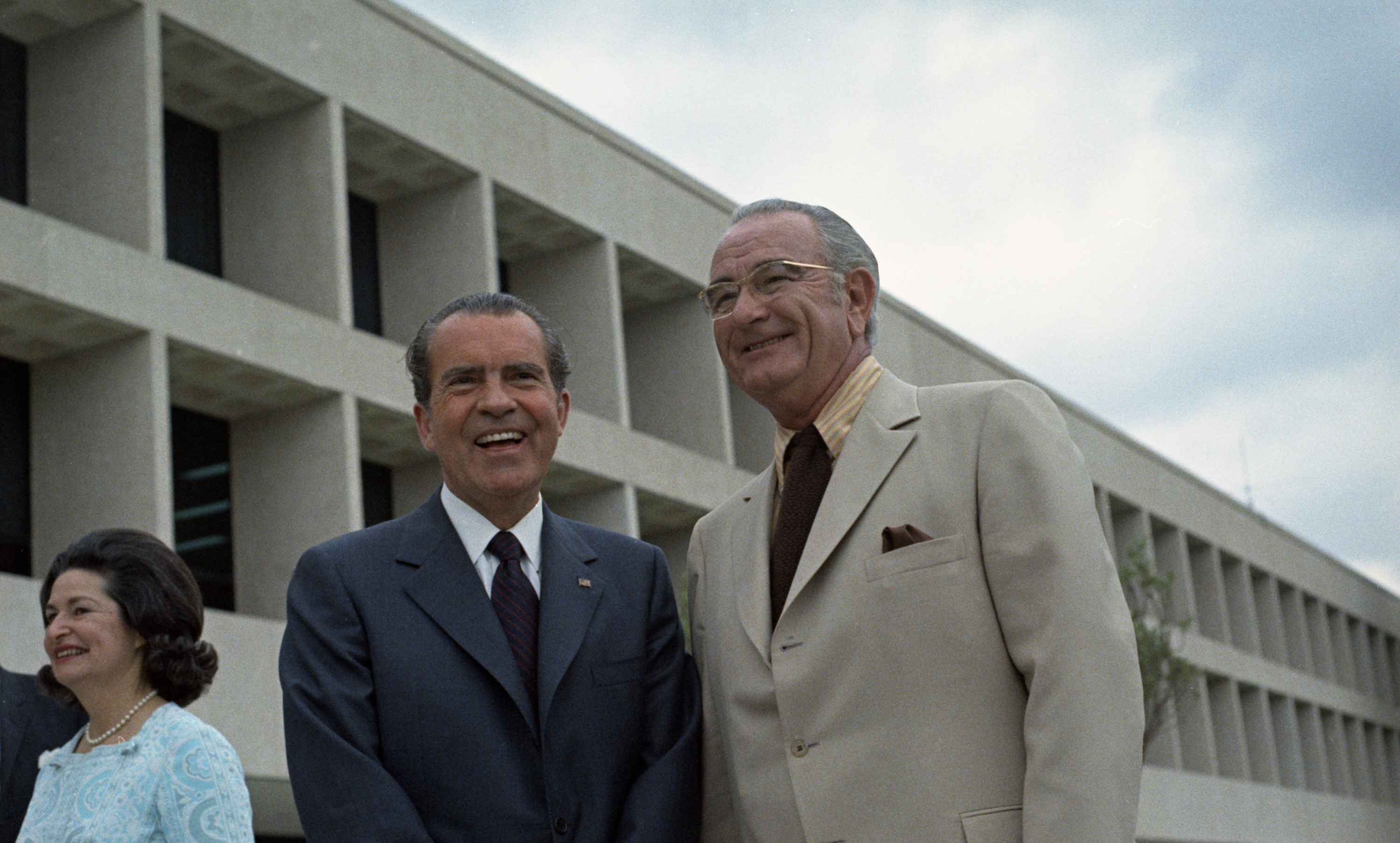 Lady Bird Johnson, Richard Nixon, and LBJ stand outside the LBJ Presidential Library with two lines of square, recessed windows tapering off to the bottom right. 